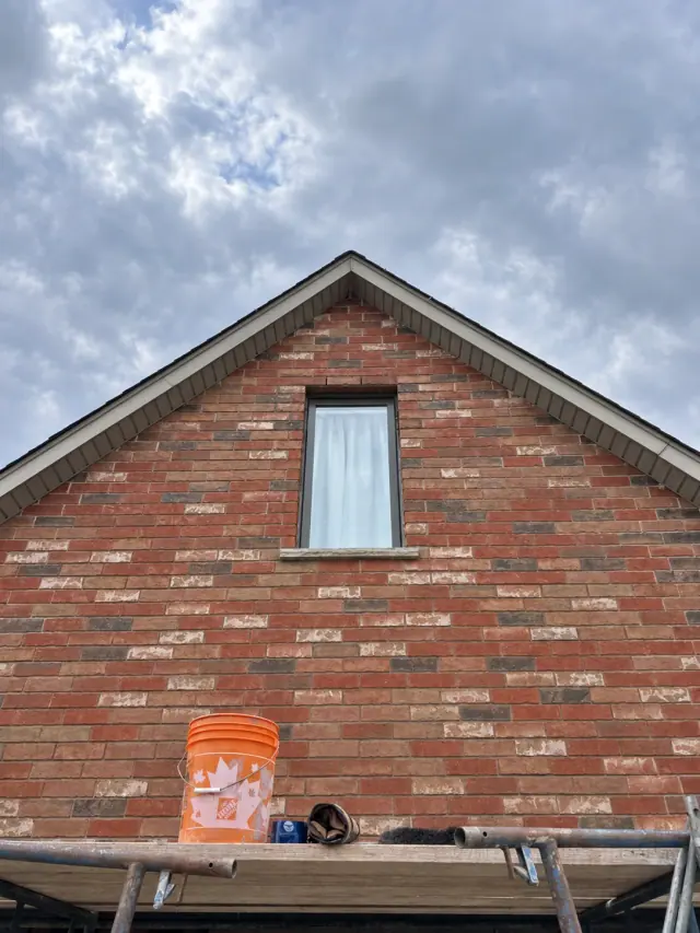 Red brick gable and narrow window above scaffolding, with a bucket and tools in the foreground