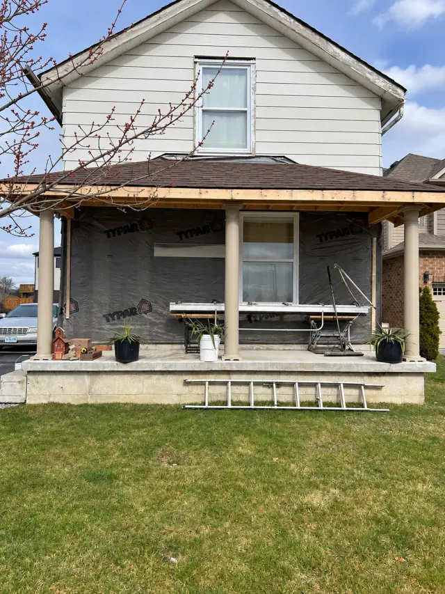 House wrap across a covered front porch before brick facing, with scaffold and a ladder on the lawn