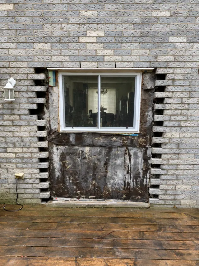 Grey brick removed beside and below a window, exposing the backing above a timber deck